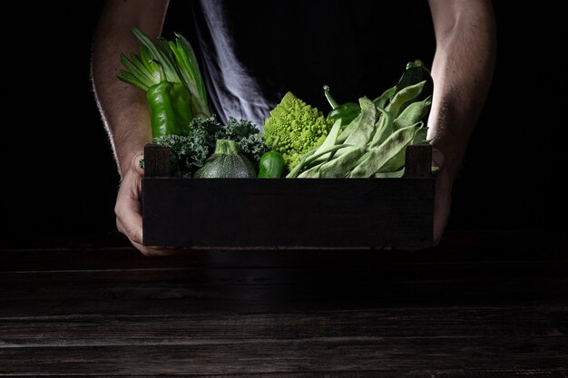 Hombre que sostiene la caja de madera con verduras sobre fondo oscuro con espacio de copia.