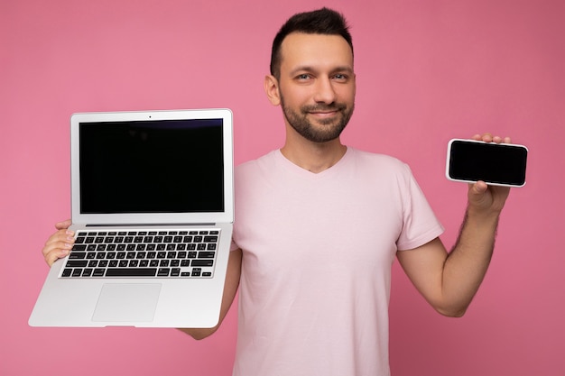 hombre que sostiene la computadora portátil y el teléfono móvil en la camiseta en la pared rosada aislada.