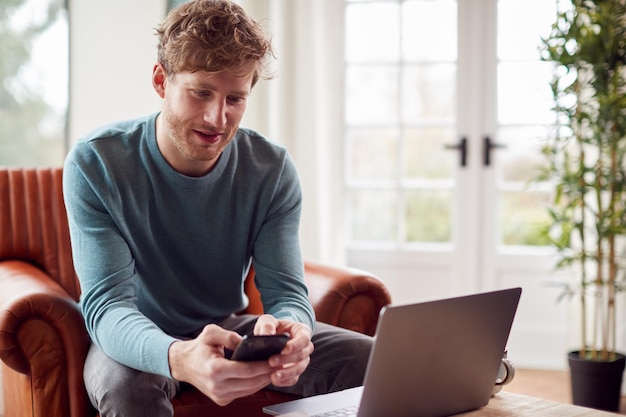 Hombre que trabaja desde casa usando una computadora portátil y un teléfono móvil