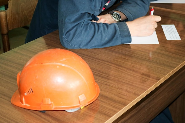 Un hombre que trabaja como ingeniero con un casco amarillo anaranjado sobre la mesa está estudiando escritura