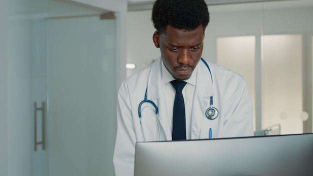 Hombre que trabaja como médico mirando la computadora para encontrar información sobre el tratamiento de la salud en la sala del hospital. Médico especialista con estetoscopio y bata blanca usando monitor para curar al paciente