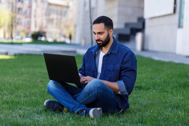 Hombre que trabaja con una computadora portátil en el césped en el parque de verano en un día brillante