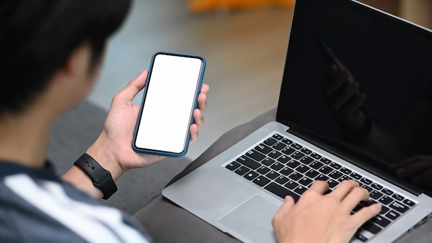 Hombre que trabaja con la computadora portátil y que usa un teléfono inteligente mientras está sentado en el sofá en casa.