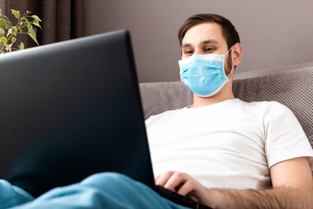 Hombre que trabaja desde la oficina en casa en máscara protectora usando laptop. Coronavirus covid 19 trabajo remoto, lugar de trabajo acogedor en el sofá