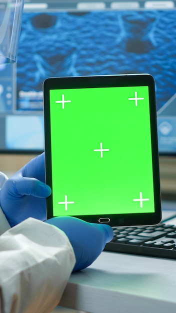 Hombre químico con traje de protección sosteniendo tableta con maqueta verde en laboratorio equipado moderno. Equipo de microbiólogos que realizan investigación de vacunas escribiendo en un dispositivo con clave de croma, pantalla aislada.