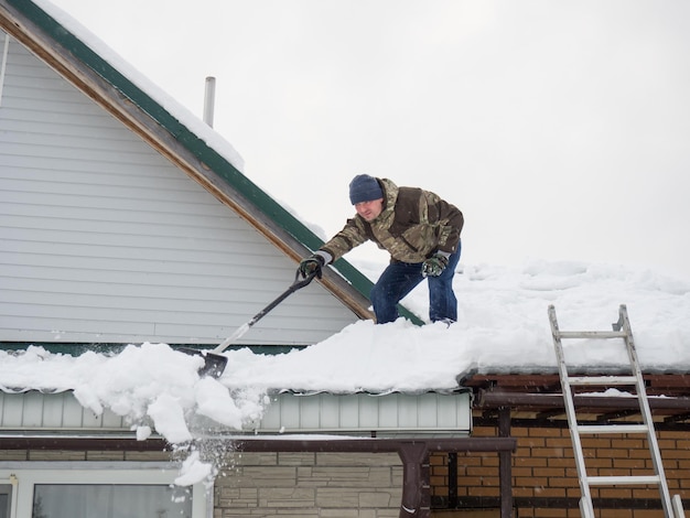 Un hombre quita la nieve del techo de una casa completamente cubierta de nieve con una pala mucha nieve fresca después de un trabajo duro y peligroso de ventisca