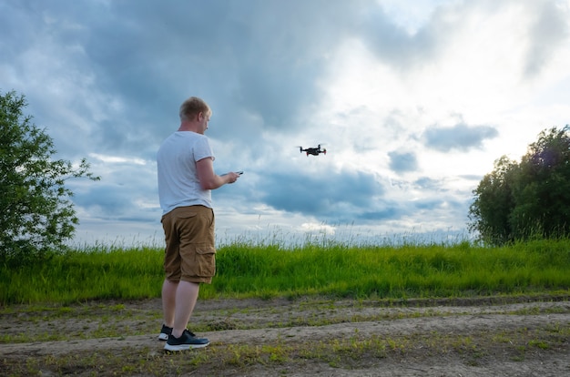 Un hombre de raza blanca con una camiseta blanca y pantalones cortos controla un quadrocopter en un claro con hierba y árboles.