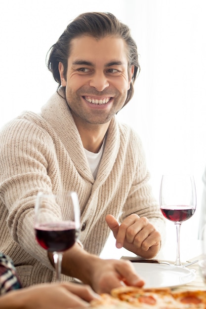 Foto hombre de raza mixta guapo positivo con rastrojo sentado a la mesa con copas de vino y charlando con amigos en la cena