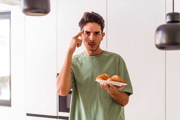 Hombre de raza mixta joven comiendo croissant en una cocina en la mañana señalando la sien con el dedo, pensando, centrado en una tarea.