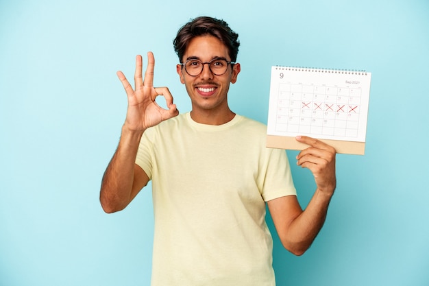 Hombre de raza mixta joven que sostiene el calendario aislado sobre fondo azul alegre y confiado que muestra el gesto ok.