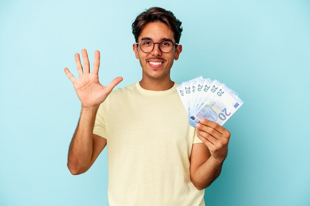 Hombre de raza mixta joven sosteniendo facturas aisladas sobre fondo azul sonriendo alegre mostrando el número cinco con los dedos.