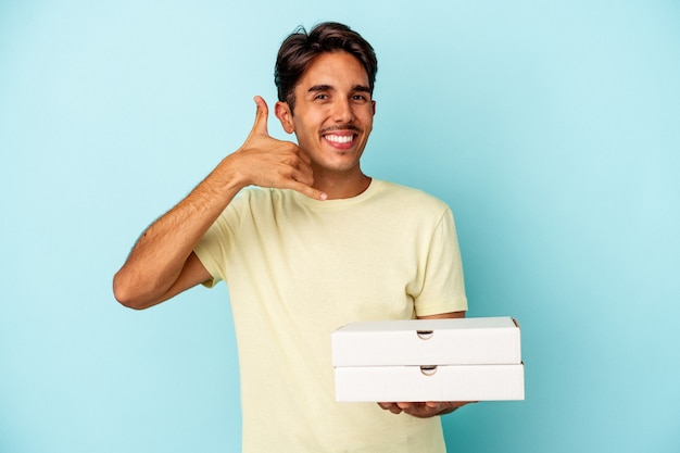 Hombre de raza mixta joven sosteniendo pizzas aisladas sobre fondo azul mostrando un gesto de llamada de teléfono móvil con los dedos.