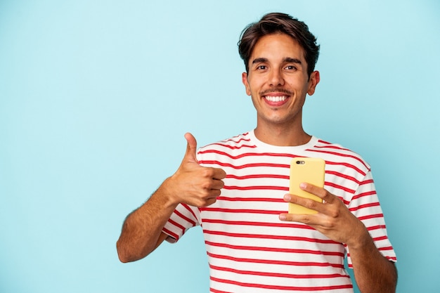 Hombre de raza mixta joven sosteniendo teléfono móvil aislado sobre fondo azul sonriendo y levantando el pulgar hacia arriba