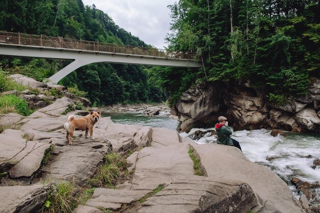 Un hombre se relaja cerca de un río de montaña mientras está sentado en una roca Un perro marrón corre cerca