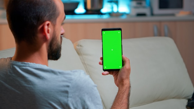 Hombre relajado mirando teléfono inteligente con simulacro de pantalla verde chroma key mientras se relaja en el sofá. Varón caucásico sosteniendo en modo horizontal teléfono con pantalla aislada a altas horas de la noche en la cocina