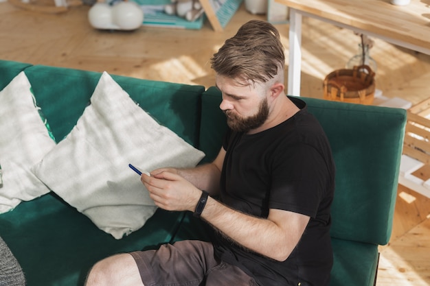 Hombre relajado usando un teléfono inteligente sentado en el sofá verde en la sala de estar en casa