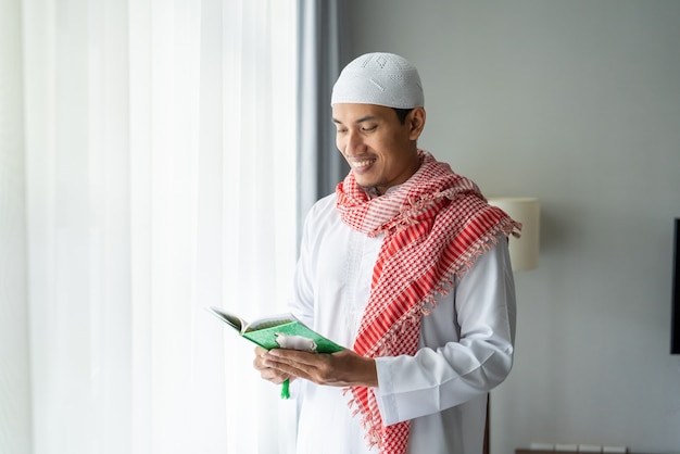 Hombre religioso asiático leyendo el Corán o el Corán mientras está de pie junto a la ventana