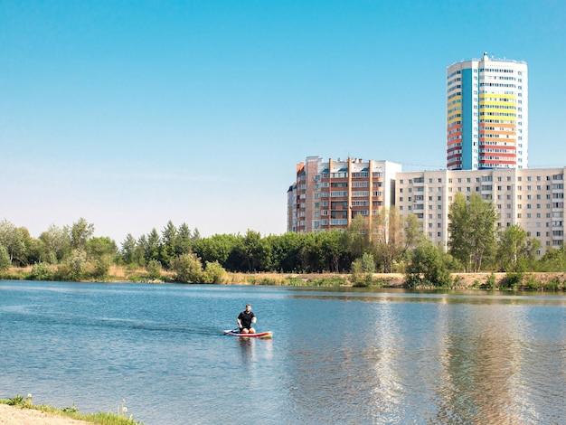 Hombre remando en paddle surf SUP en el lago de la ciudad entre las casas