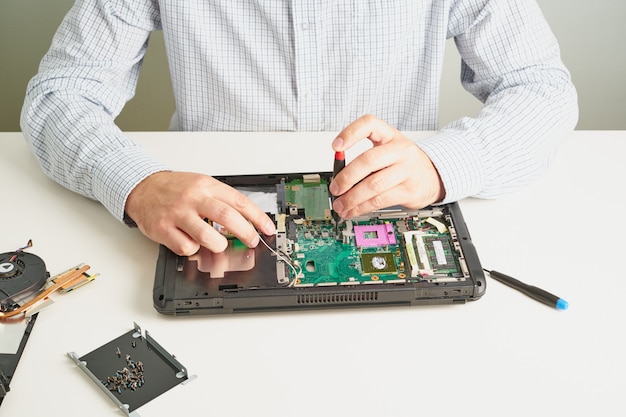 El hombre repara la computadora. Un ingeniero de servicio en camisa reparaciones portátil, en blanco Escritorio contra la pared blanca.