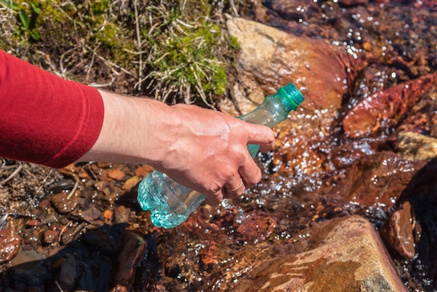 El hombre repone el agua del arroyo de la montaña bajo el sol brillante El turista llena la botella con agua mineral en un día soleado Mano con la botella en el arroyo de agua clara a la luz del sol Reposición de agua durante la caminata