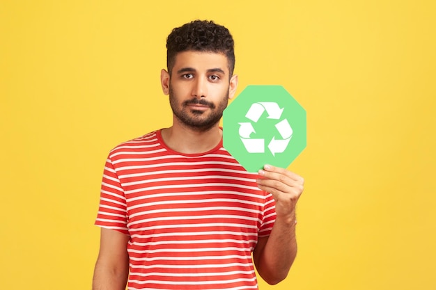 Hombre responsable y seguro de sí mismo con barba en camiseta a rayas sosteniendo un cartel de reciclaje verde en la mano y mirando seriamente a la cámara pensando en una toma de estudio interior verde aislada en un fondo amarillo