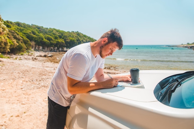 Hombre revisando el mapa en el lugar de destino del capó del automóvil playa de mar en el fondo