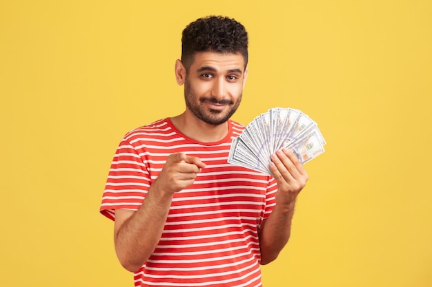 Hombre rico y seguro de sí mismo con barba en camiseta a rayas apuntándote con el dedo sosteniendo un abanico de billetes en dólares sugiriendo ganar jack pot Estudio interior aislado en fondo amarillo