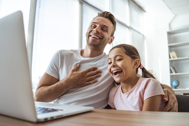 El hombre riendo y una niña con una computadora portátil se sientan en el escritorio