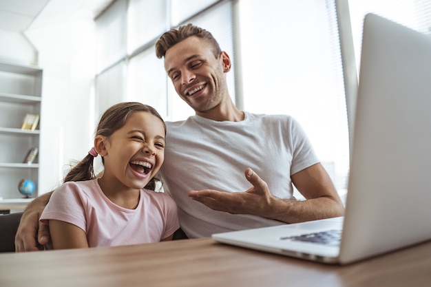 El hombre riendo y una niña con una computadora portátil se sientan en el escritorio