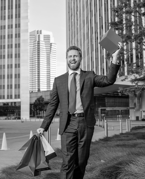 Foto hombre riendo en ropa formal con bolsas de compras y caja de regalo camina fuera del saludo de la oficina