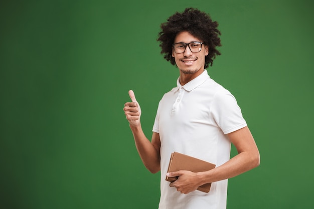 Hombre rizado africano joven feliz que sostiene el libro que muestra los pulgares para arriba.