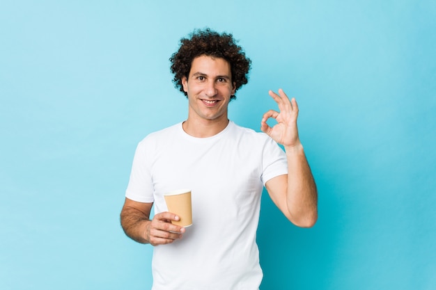 Hombre rizado caucásico joven que sostiene un café para llevar alegre y confiado que muestra gesto aceptable.