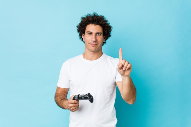 Hombre rizado caucásico joven que sostiene un controlador de juego que muestra el número uno con el dedo.