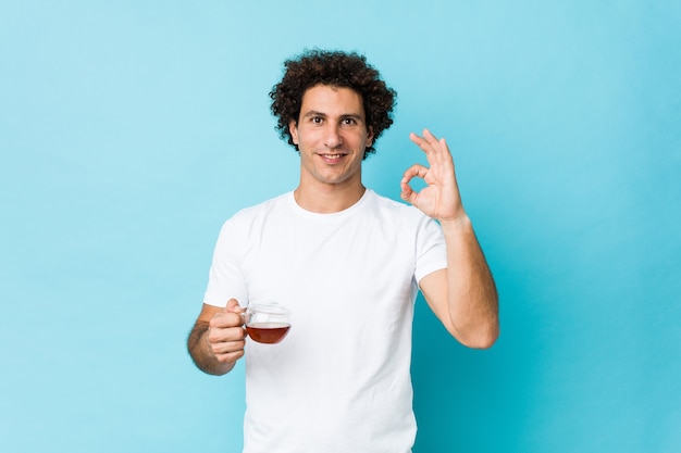 Foto hombre rizado caucásico joven que sostiene una taza de té alegre y confiado que muestra el gesto aceptable
