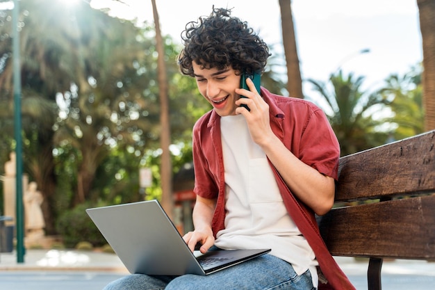 Hombre rizado caucásico joven que trabaja usando la computadora portátil y hablando en el teléfono inteligente