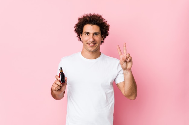 Hombre rizado caucásico joven sosteniendo un vape mostrando el signo de la victoria y sonriendo ampliamente.