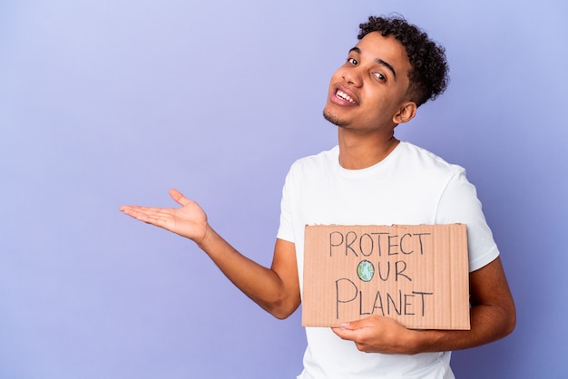 Hombre rizado joven aislado sosteniendo un proteger nuestro planeta mostrando un espacio de copia en una palma y sosteniendo otra mano en la cintura