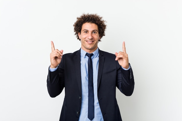 El hombre rizado joven del negocio contra la pared blanca indica con ambos dedos delanteros para arriba que muestran un espacio en blanco.
