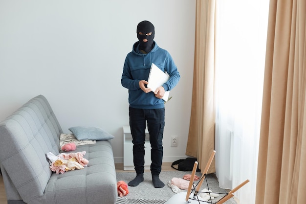 El hombre roba una computadora portátil en la casa de otra persona huye siendo perseguido usa una máscara de ladrón y una sudadera con capucha azul parado cerca del sofá y la ventana robo peligroso rompiendo plano