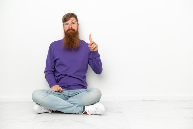 Hombre rojizo caucásico joven sentado en el suelo aislado sobre fondo blanco señalando con el dedo índice una gran idea