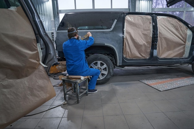 Hombre con ropa azul protectora sentado cerca del auto y haciendo molienda de carrocerías antes de pintar e imprimar