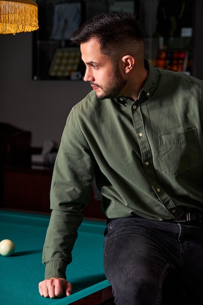 Hombre en ropa casual se sienta en la mesa de billar, mirando el juego, concepto de juego de deporte de billar. retrato