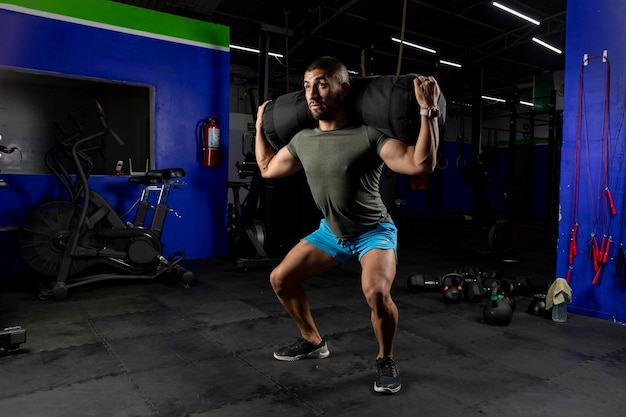 Hombre en ropa deportiva en un gimnasio haciendo sentadillas con un saco de arena en la espalda