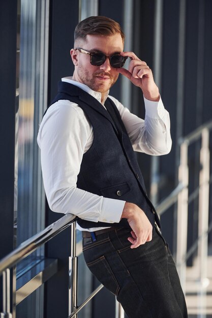 Foto el hombre con ropa elegante y gafas de sol está parado adentro y posando para la cámara.