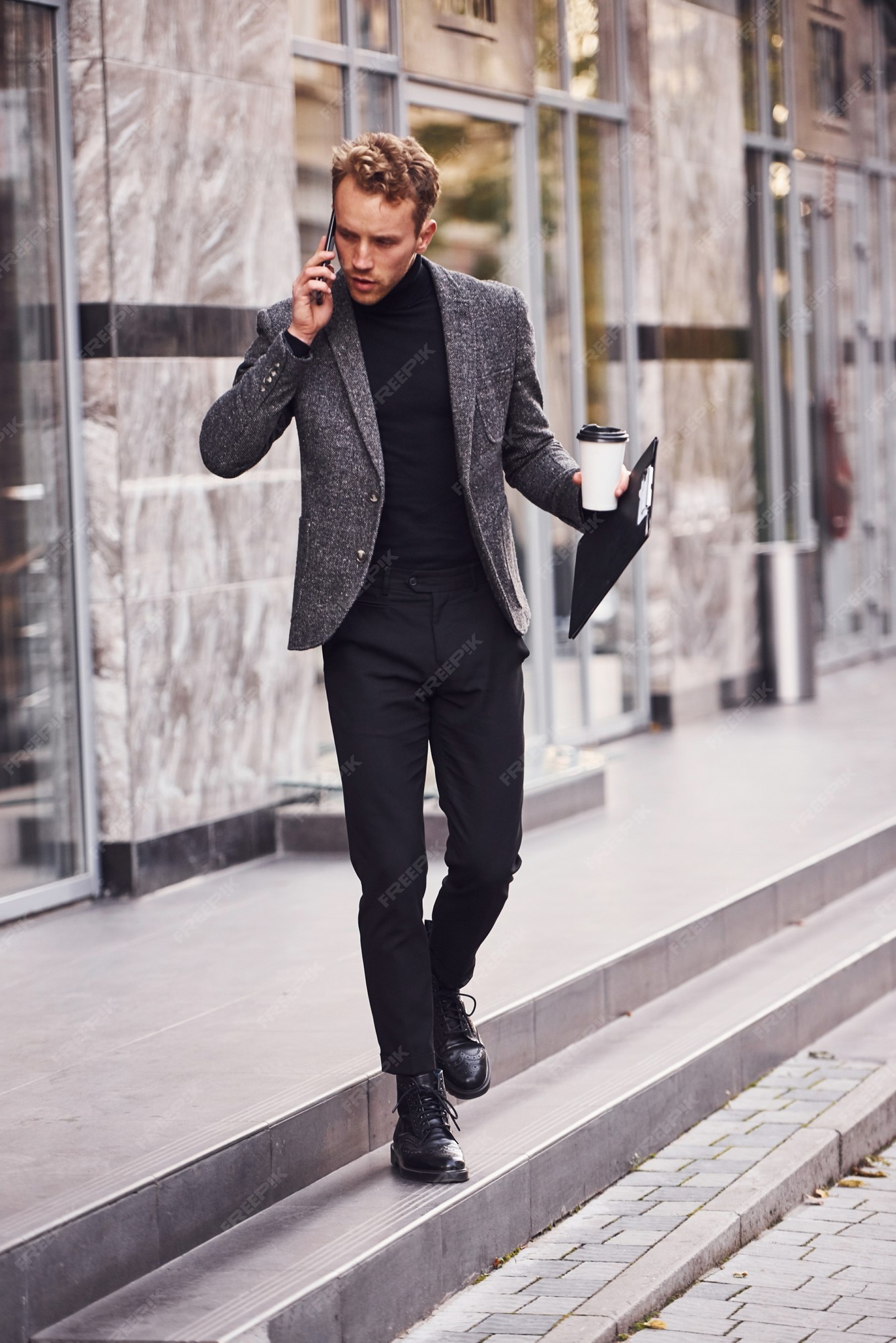 El hombre en formal elegante con una taza de bebida y un bloc de notas en las manos está afuera contra el edificio moderno y tiene una conversación por teléfono.