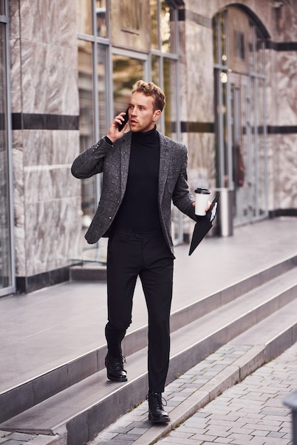 El hombre en formal elegante con una taza de bebida y un bloc de notas en las manos está afuera contra el edificio moderno y tiene una conversación por teléfono.