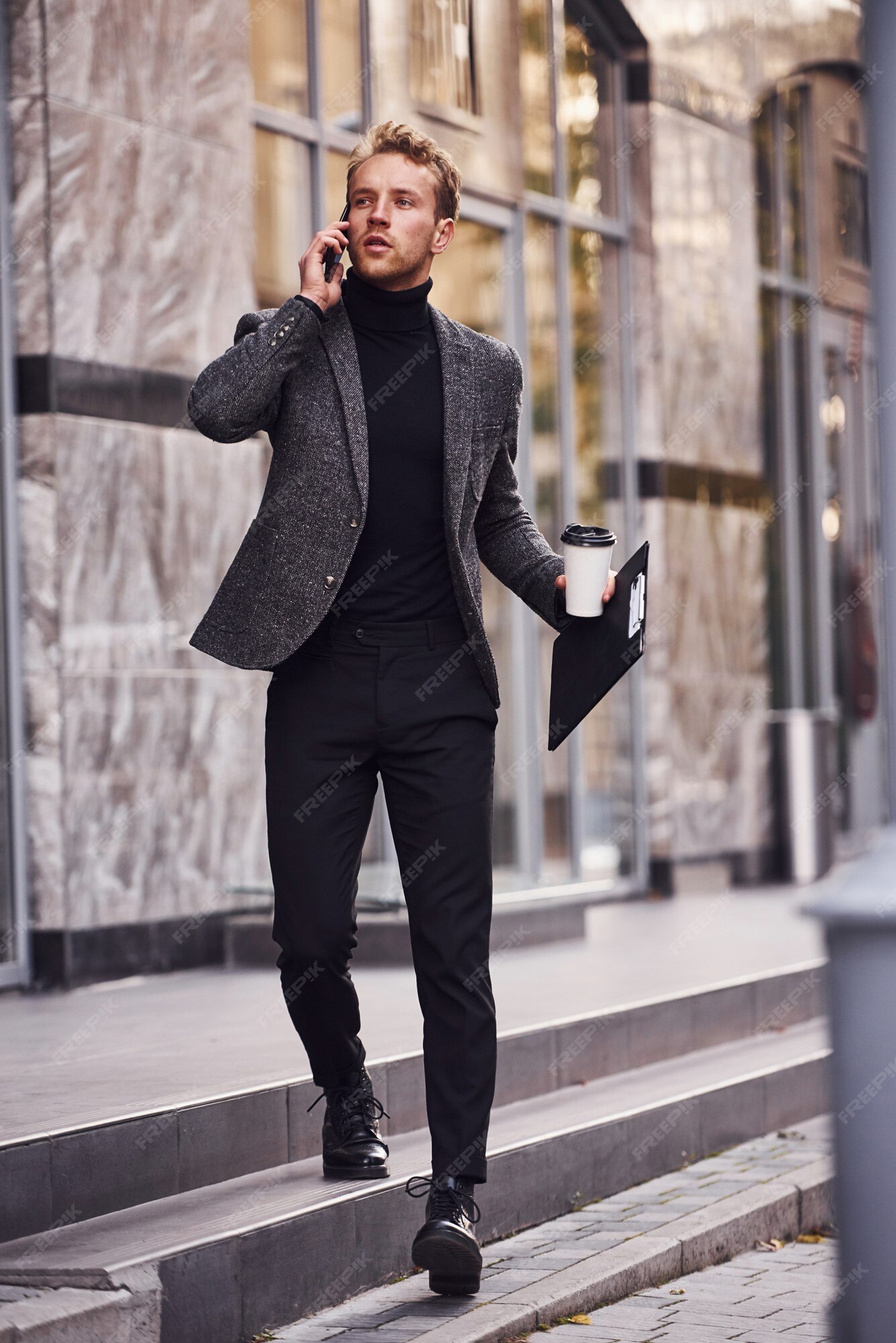 El hombre en ropa formal elegante con una taza de bebida y un bloc de notas en manos está afuera contra el edificio moderno y una conversación por teléfono.