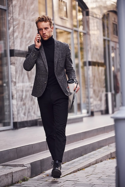 El hombre en formal elegante con una taza de bebida y un bloc de notas en las manos está afuera contra el edificio moderno y tiene una conversación por teléfono.