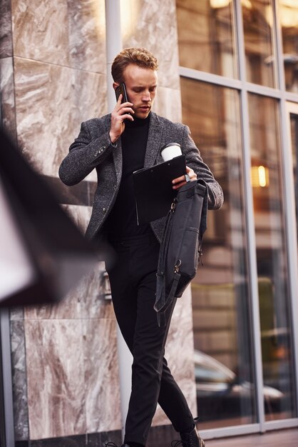 El hombre en ropa formal elegante con una taza de bebida y un bloc de notas en las manos está afuera contra el edificio moderno y tiene una conversación por teléfono.