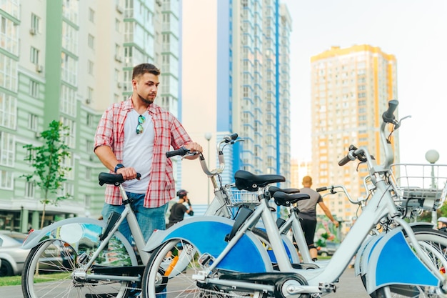 El hombre con ropa informal toma una bicicleta del estacionamiento en un paseo nocturno. El tipo con barba alquila una bicicleta en el fondo del paisaje urbano. Bicicleta compartida.